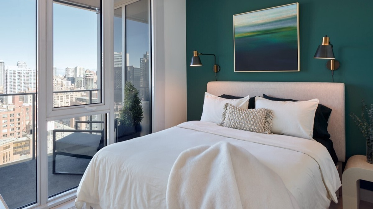 Modern bedroom with deep green accent wall, upholstered bed, brass sconces, and balcony skyline views at 369 Grand apartments in Chicago