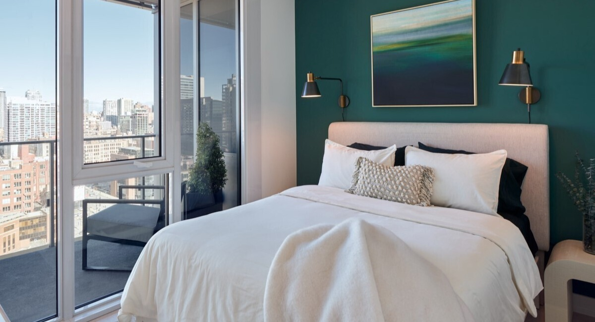 Modern bedroom with deep green accent wall, upholstered bed, brass sconces, and balcony skyline views at 369 Grand apartments in Chicago