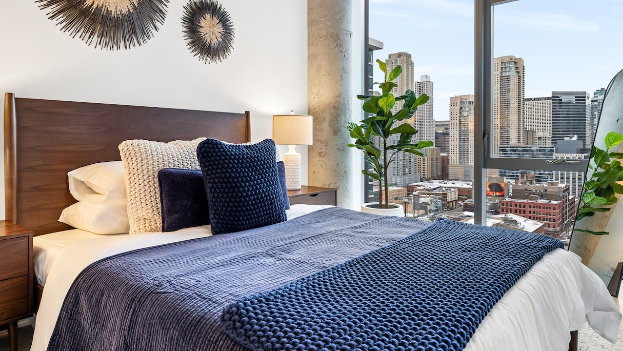 Cozy bedroom at 3Eleven Luxury Apartments in Chicago featuring blue bedding, wood headboard, warm lamps, and large windows with skyline views