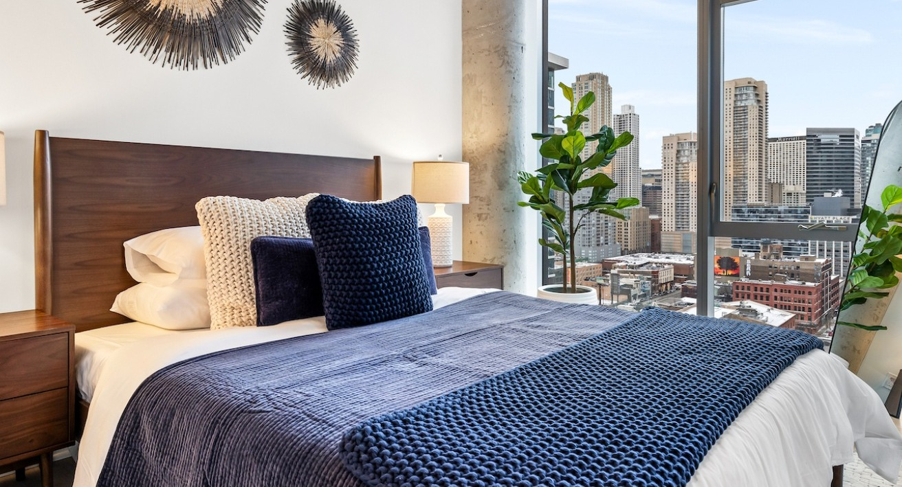 Cozy bedroom at 3Eleven Luxury Apartments in Chicago featuring blue bedding, wood headboard, warm lamps, and large windows with skyline views