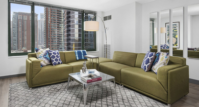 Sunlit living room with twin green sofas, arc floor lamp, and expansive windows showcasing Chicago skyline views at 420 East Ohio apartments—ideal for relaxing or entertaining