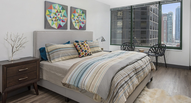 Cozy bedroom with striped bedding, colorful geometric wall art, large windows, and city views at 420 East Ohio apartments in Chicago; wood floors and a soft rug add warmth