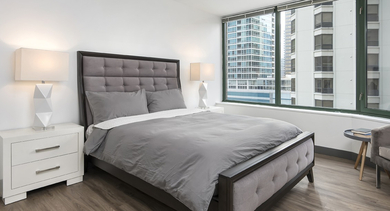 Spacious primary bedroom with tufted gray bed, matching white nightstands, and sweeping bay windows at 420 East Ohio apartments in Chicago, bringing in generous natural light