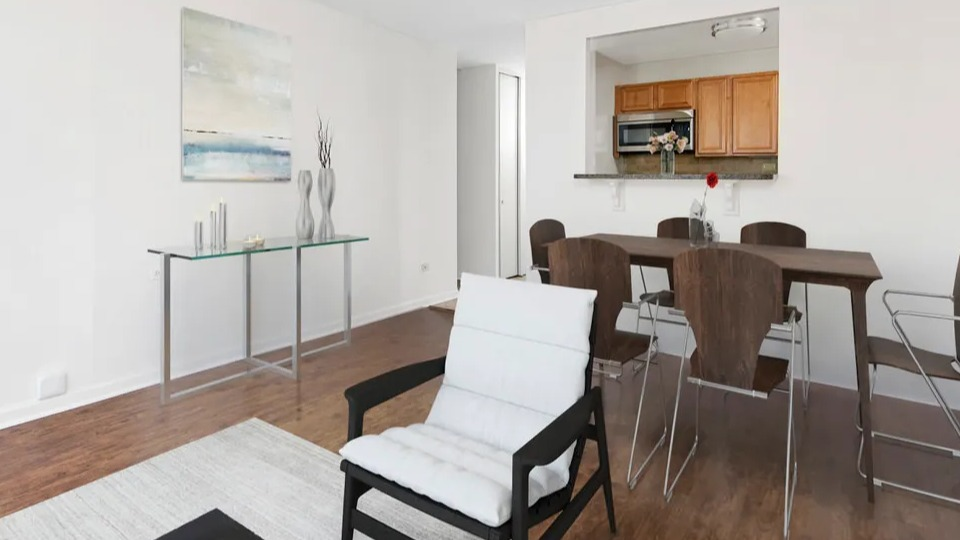 Open living and dining area with white accent chair, glass console, six-seat table, and pass-through kitchen at 55 West Chestnut apartments in Chicago
