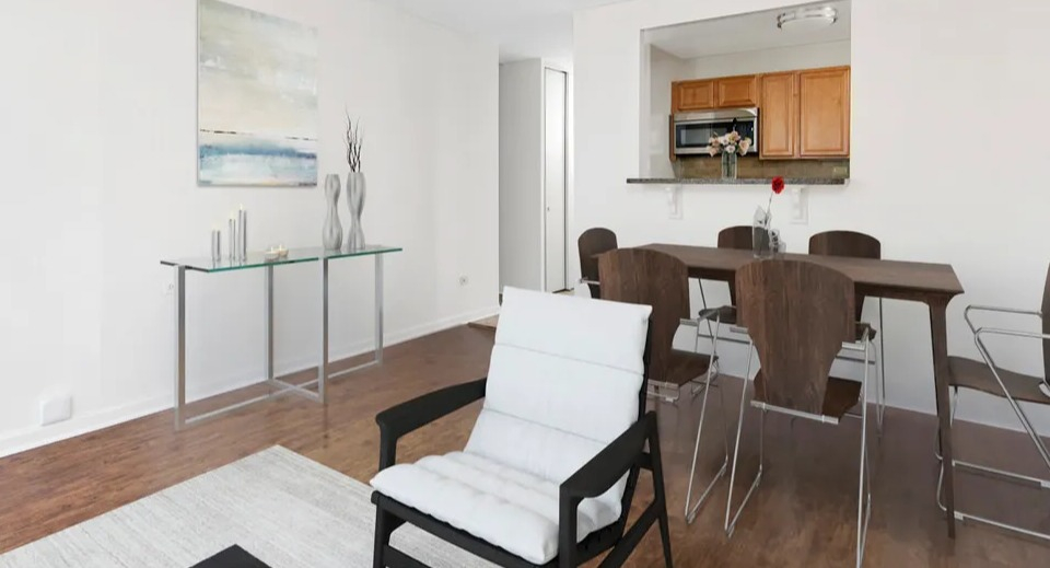 Open living and dining area with white accent chair, glass console, six-seat table, and pass-through kitchen at 55 West Chestnut apartments in Chicago