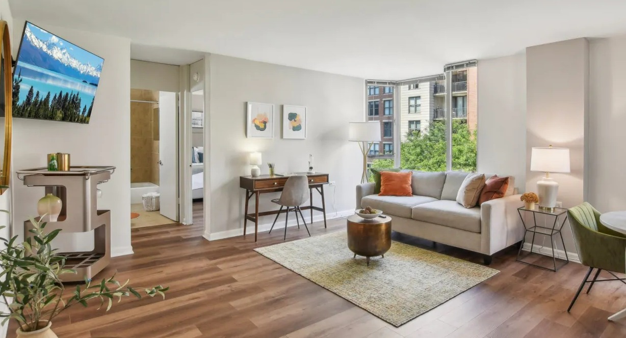 Bright open-plan living area with sofa, work desk, round dining set, and wood floors with tree-lined views at 55 West Chestnut apartments in Chicago