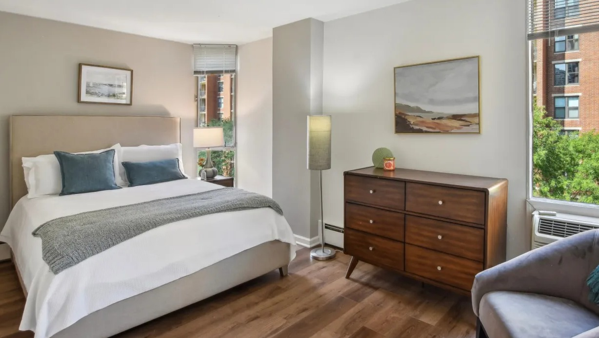 Peaceful bedroom with king bed, white bedding, mid-century dresser, accent chair, and large windows at 55 West Chestnut apartments in Chicago
