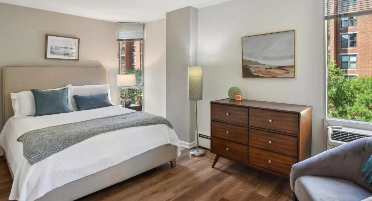 Peaceful bedroom with king bed, white bedding, mid-century dresser, accent chair, and large windows at 55 West Chestnut apartments in Chicago