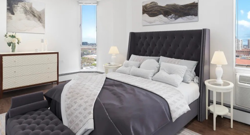 Stylish bedroom with tufted headboard, upholstered bench, bedside tables, and two tall windows bringing skyline views at 55 West Chestnut apartments in Chicago