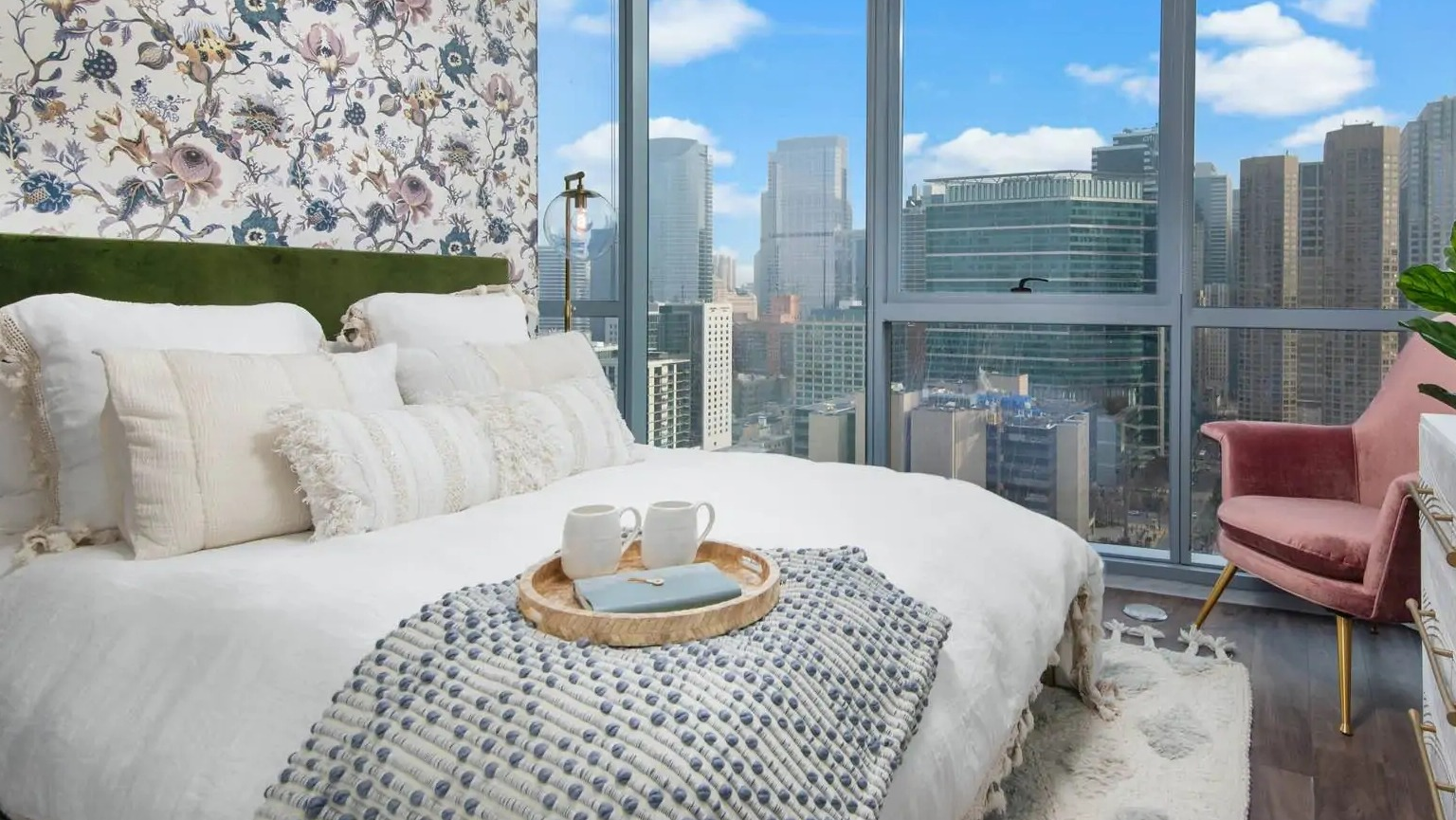 Cozy bedroom at 727 West Madison in Chicago with floor-to-ceiling windows, skyline views, floral accent wall, and layered bedding