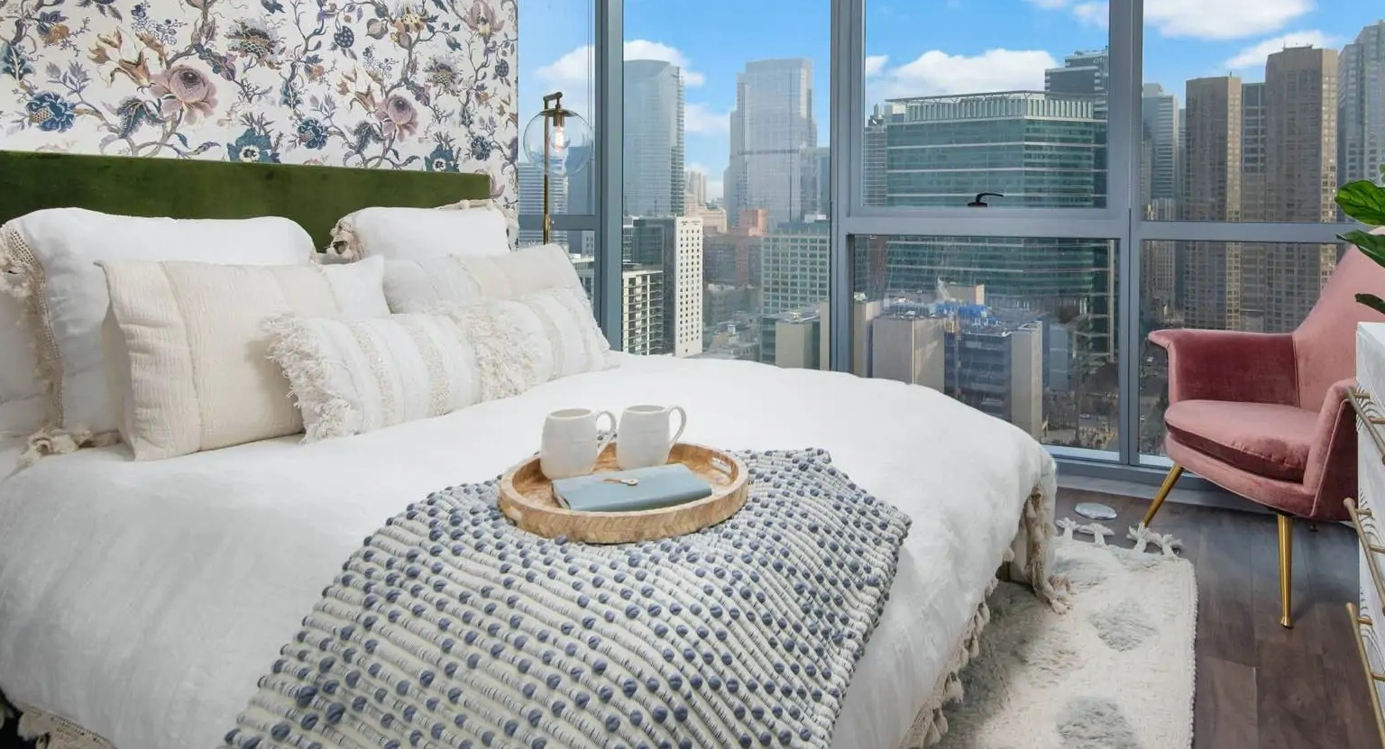 Cozy bedroom at 727 West Madison in Chicago with floor-to-ceiling windows, skyline views, floral accent wall, and layered bedding