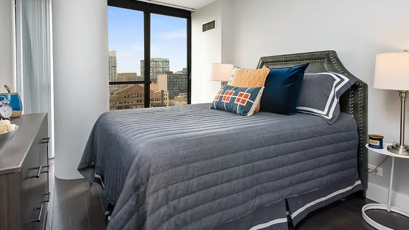 Cozy bedroom with corner windows, upholstered headboard, and layered gray bedding at 777 South State apartments in Chicago