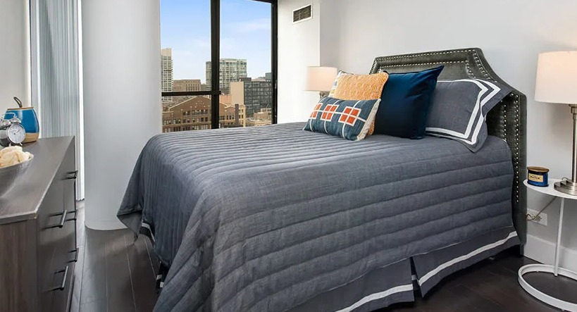 Cozy bedroom with corner windows, upholstered headboard, and layered gray bedding at 777 South State apartments in Chicago