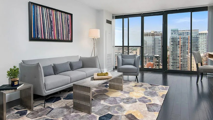 Bright living room with floor-to-ceiling windows, skyline views, dark wood floors, and modern gray seating at 777 South State apartments in Chicago