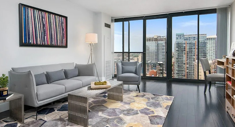 Bright living room with floor-to-ceiling windows, skyline views, dark wood floors, and modern gray seating at 777 South State apartments in Chicago