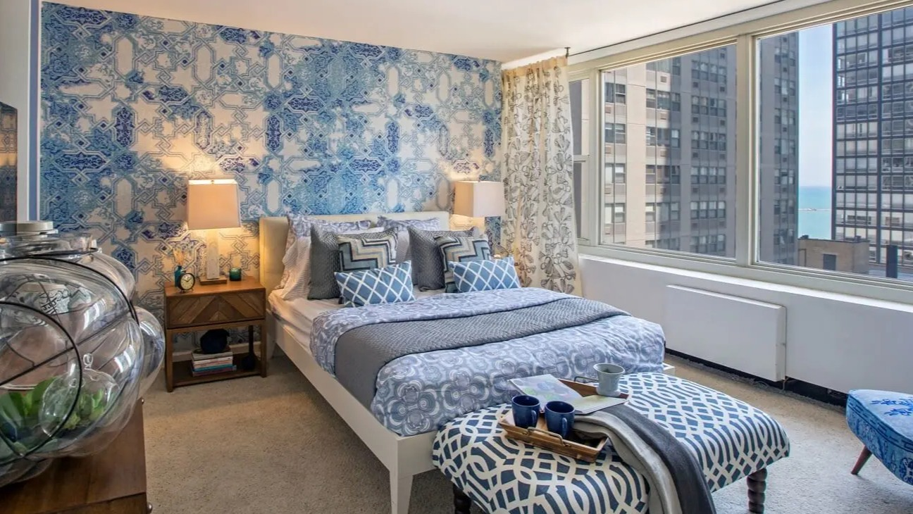 Sun-filled bedroom with large windows, blue accent wall, plush carpet, and city and lake views at 860 N Dewitt Apartments in Chicago