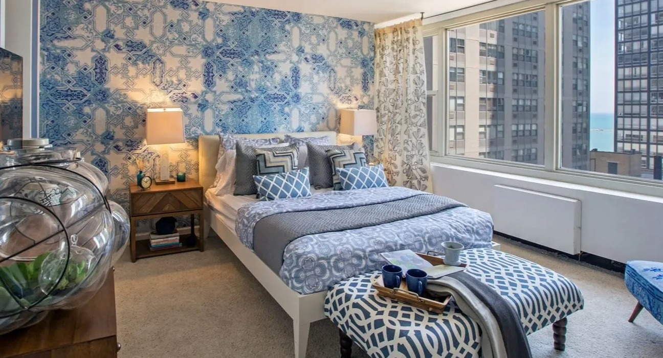 Sun-filled bedroom with large windows, blue accent wall, plush carpet, and city and lake views at 860 N Dewitt Apartments in Chicago