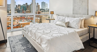 Bright bedroom with floor-to-ceiling windows, upholstered bed, bedside lamps, and sweeping city skyline views at Aberdeen East Apartments in Chicago