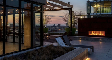 Rooftop lounge and terrace with glass-walled clubhouse, pergola string lights, and linear fireplace overlooking the city at Aberdeen East Apartments in Chicago