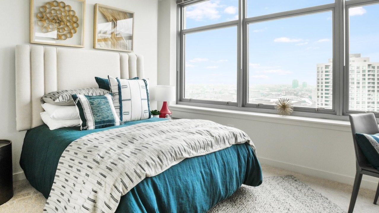 Sunlit bedroom at Alta at K Station showcasing expansive windows, Chicago skyline views, an upholstered headboard, and layered bedding in modern tones