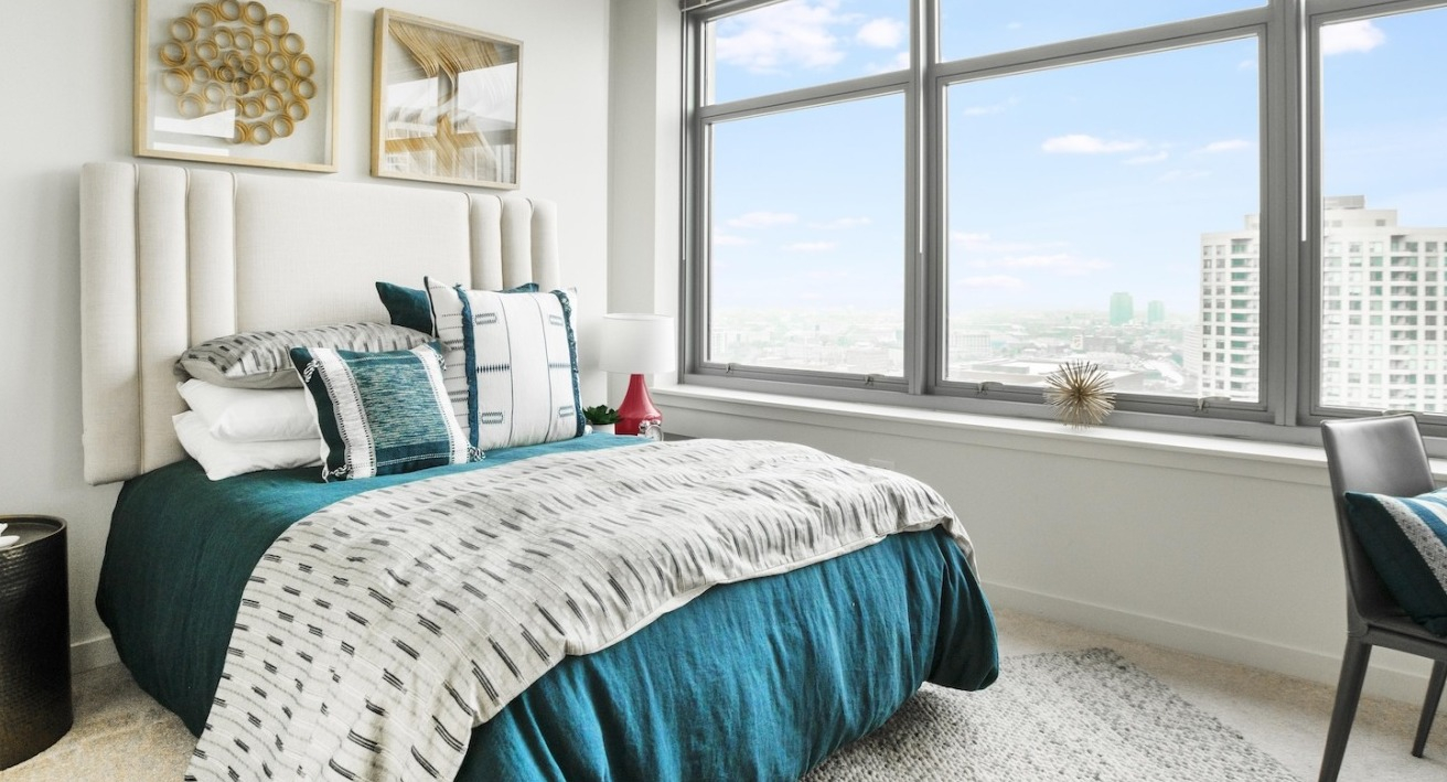 Sunlit bedroom at Alta at K Station showcasing expansive windows, Chicago skyline views, an upholstered headboard, and layered bedding in modern tones