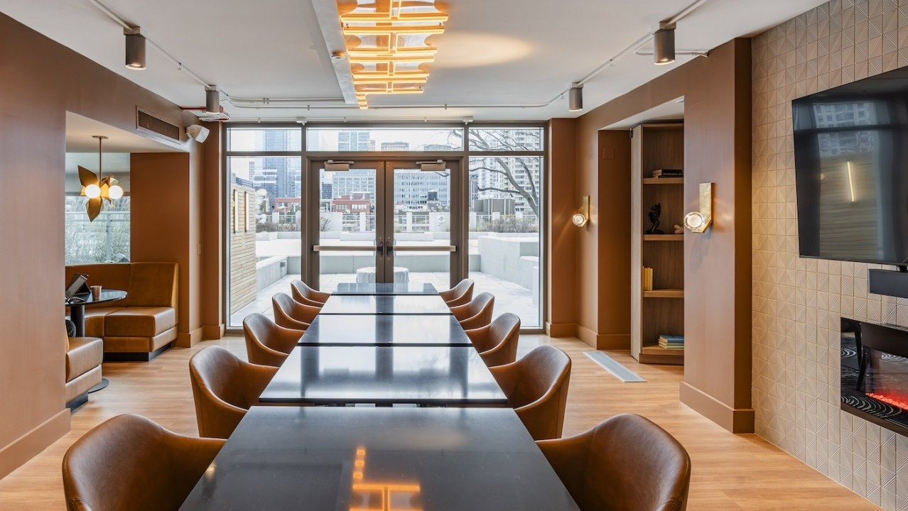 Coworking lounge at Alta at K Station with black tables, tan seating, wall-mounted TV, fireplace, and private booths along the side