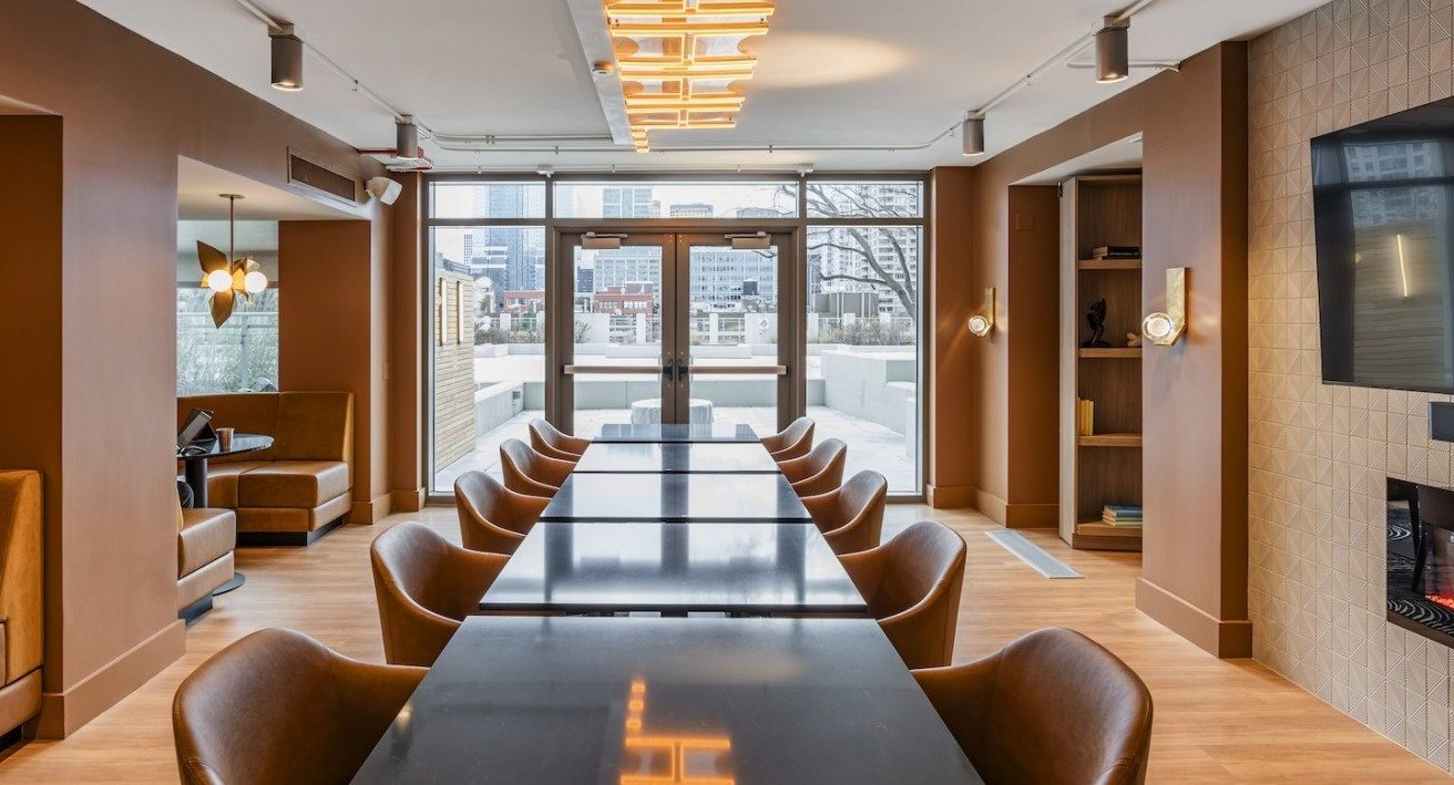 Coworking lounge at Alta at K Station with black tables, tan seating, wall-mounted TV, fireplace, and private booths along the side