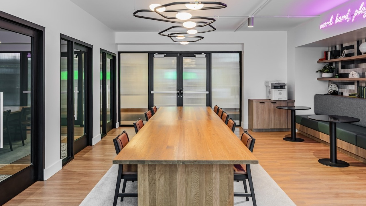Bright coworking room at Alta at K Station with long communal table, booth seating, glass-partition offices, and neon accent lighting