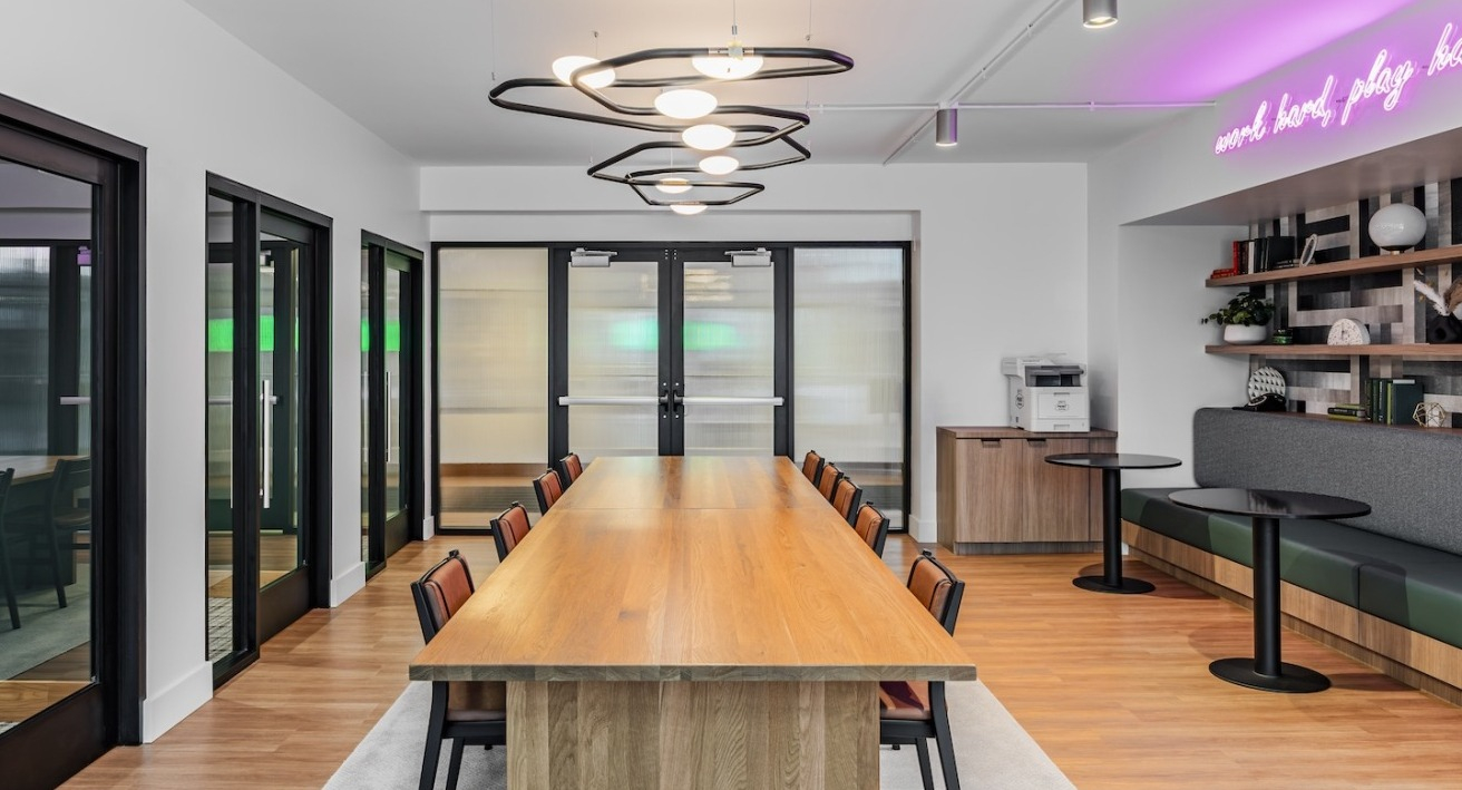 Bright coworking room at Alta at K Station with long communal table, booth seating, glass-partition offices, and neon accent lighting