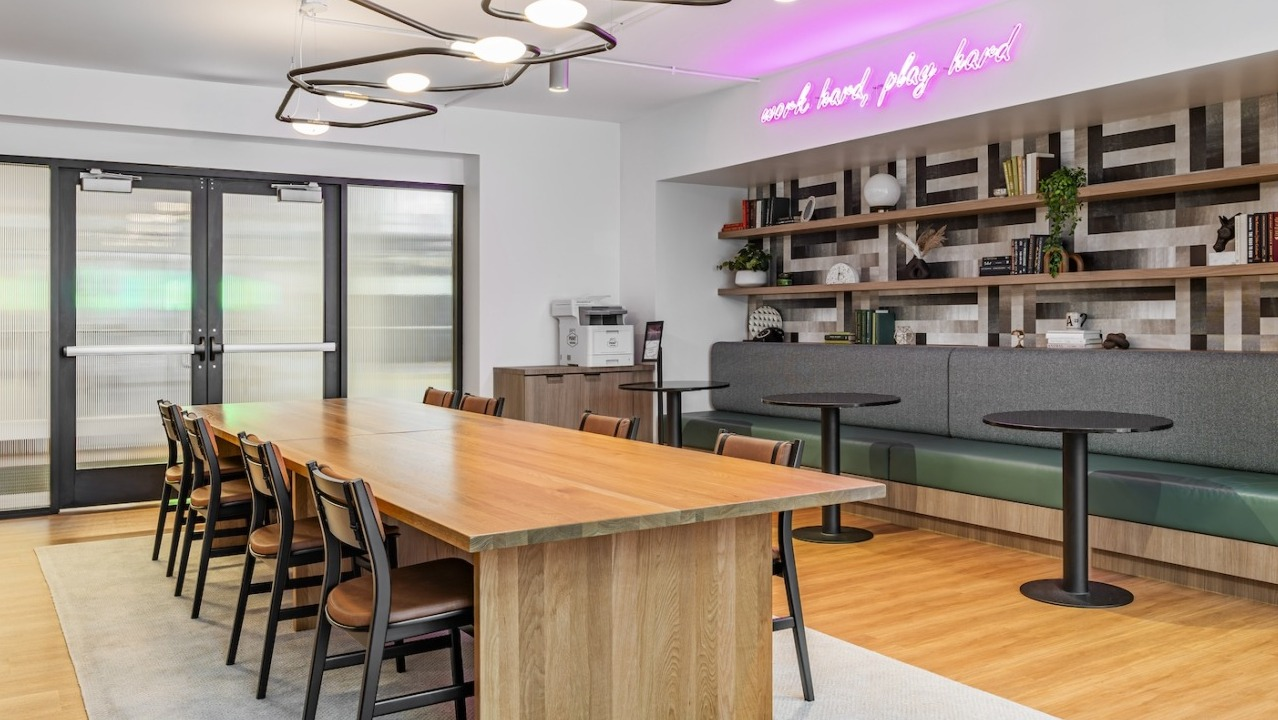 Modern coworking space at Alta at K Station featuring long wooden table, library shelves, booth seating, and neon “work hard play hard” sign
