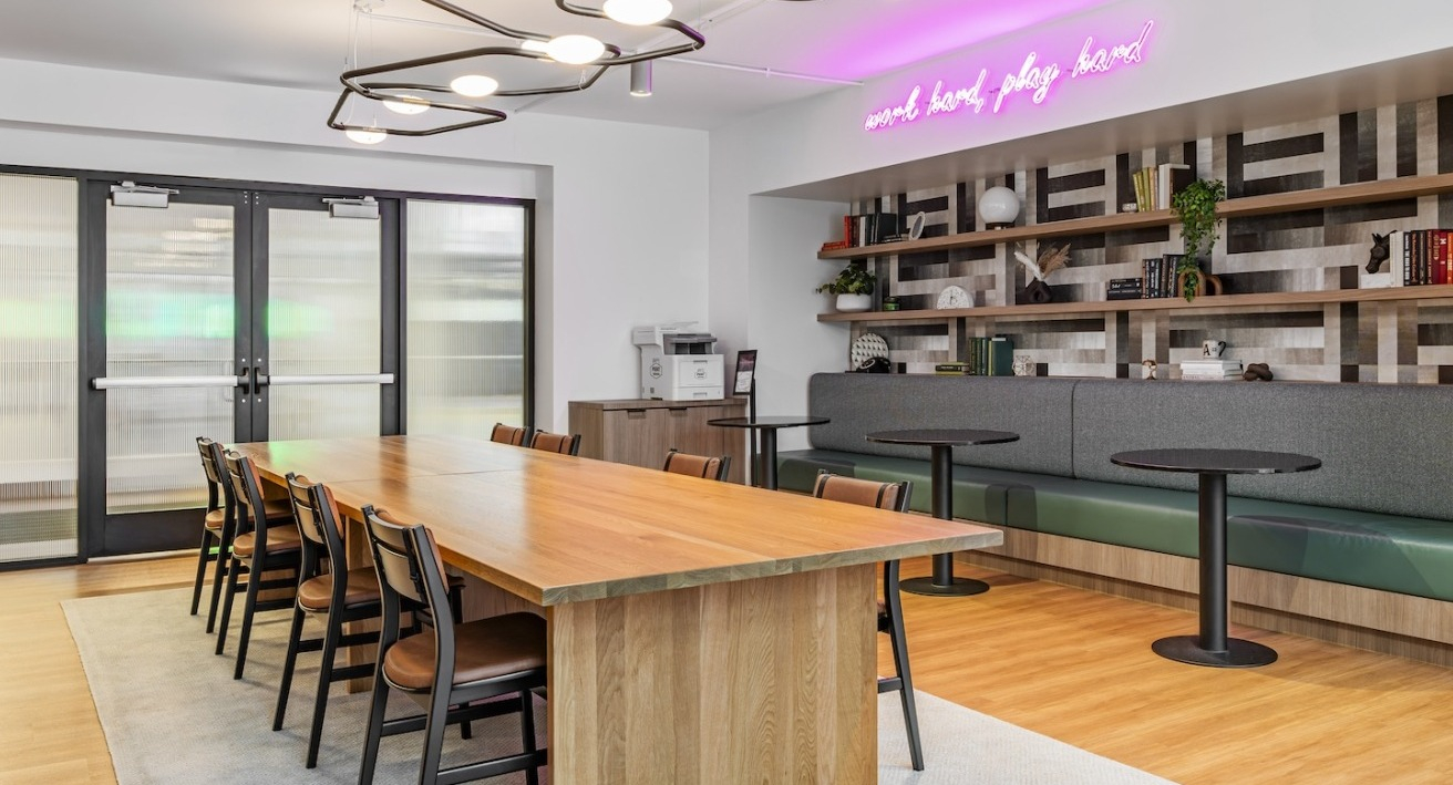 Modern coworking space at Alta at K Station featuring long wooden table, library shelves, booth seating, and neon “work hard play hard” sign
