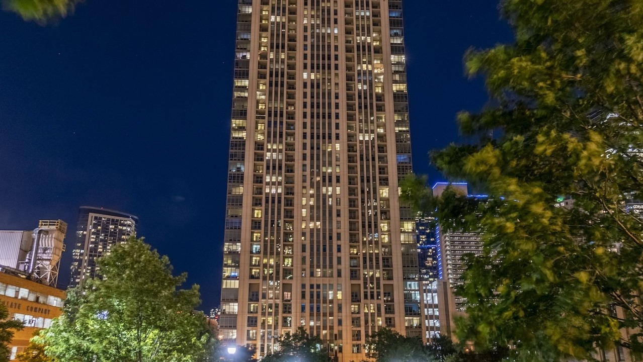 Exterior night view of Alta at K Station tower in Chicago, with illuminated windows, city skyline backdrop, and trees in the foreground