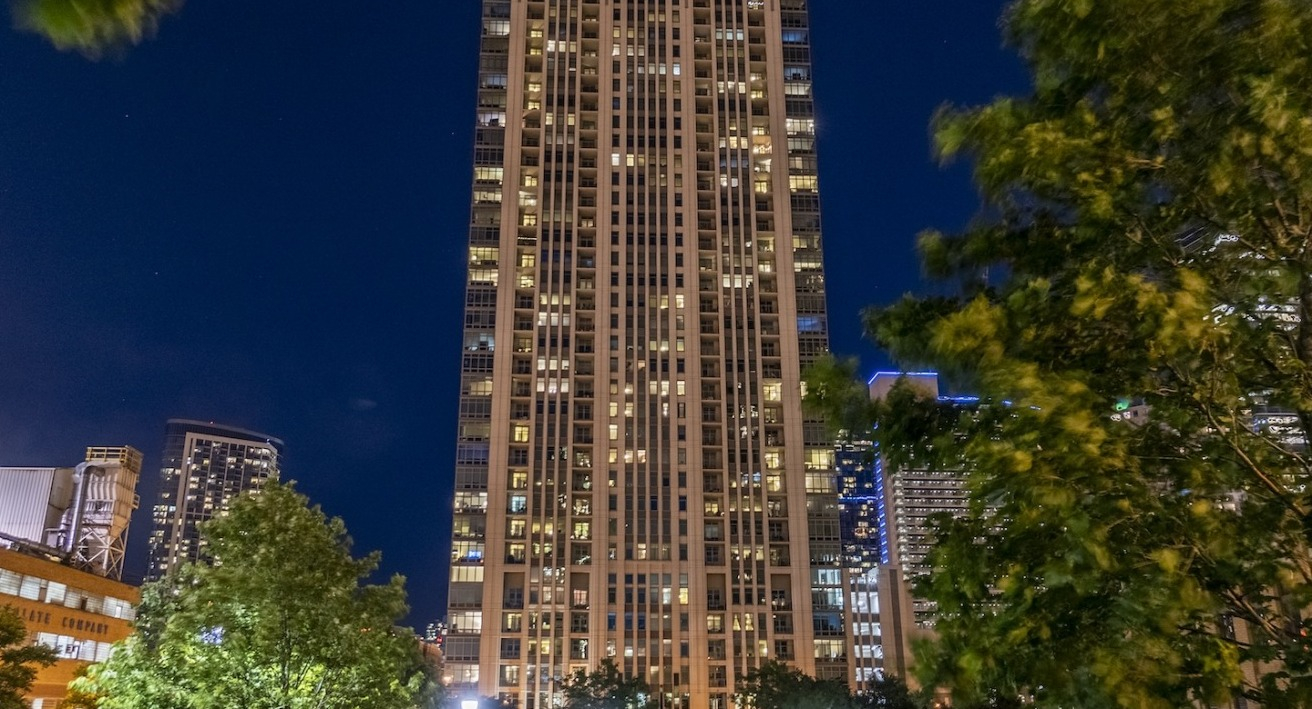 Exterior night view of Alta at K Station tower in Chicago, with illuminated windows, city skyline backdrop, and trees in the foreground