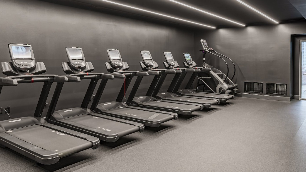 Cardio fitness room at Alta at K Station featuring multiple treadmills, stair machine, dark modern interior, and access to outdoor patio
