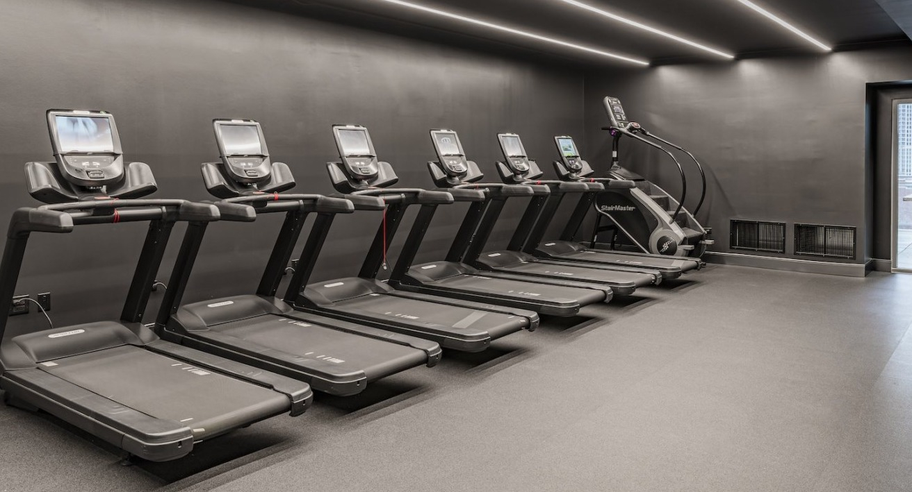 Cardio fitness room at Alta at K Station featuring multiple treadmills, stair machine, dark modern interior, and access to outdoor patio
