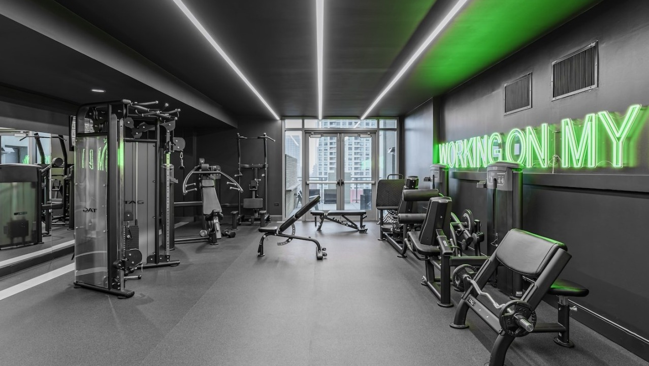 Strength training gym at Alta at K Station with weight machines, benches, mirrored wall, and neon green “working on my fit” wall décor