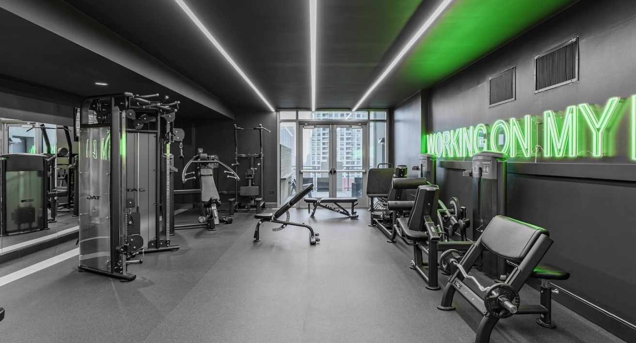 Strength training gym at Alta at K Station with weight machines, benches, mirrored wall, and neon green “working on my fit” wall décor