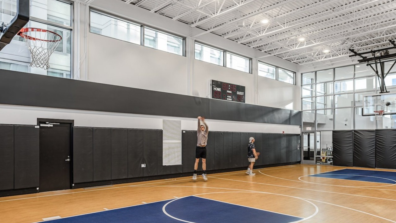 Indoor basketball court at Alta at K Station with full-size hoops, wood flooring, scoreboard, and natural light from floor-to-ceiling windows