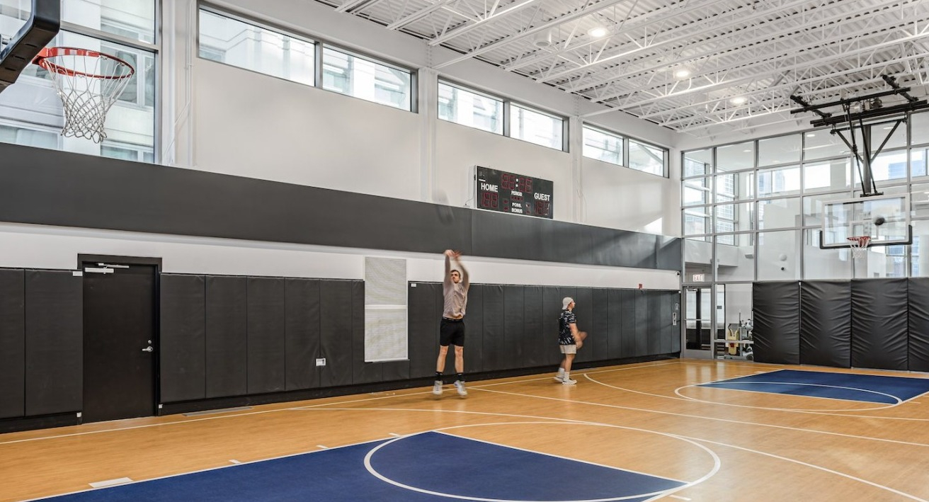 Indoor basketball court at Alta at K Station with full-size hoops, wood flooring, scoreboard, and natural light from floor-to-ceiling windows
