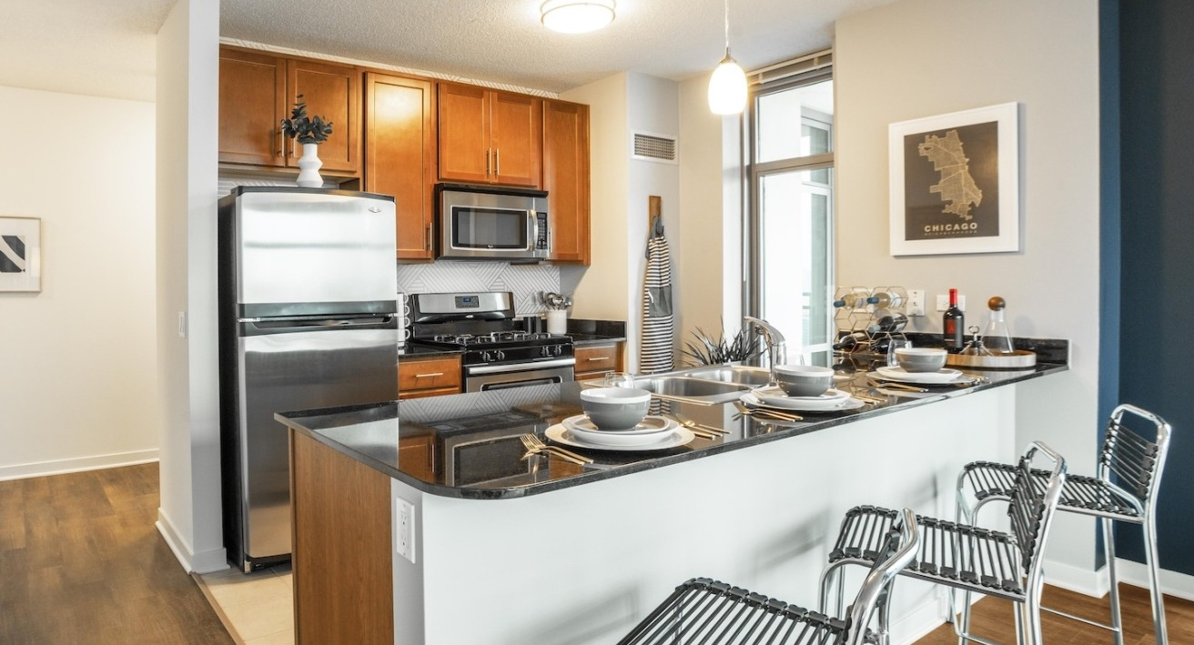Modern kitchen at Alta at K Station with stainless steel appliances, wood cabinetry, granite countertops, and breakfast bar seating