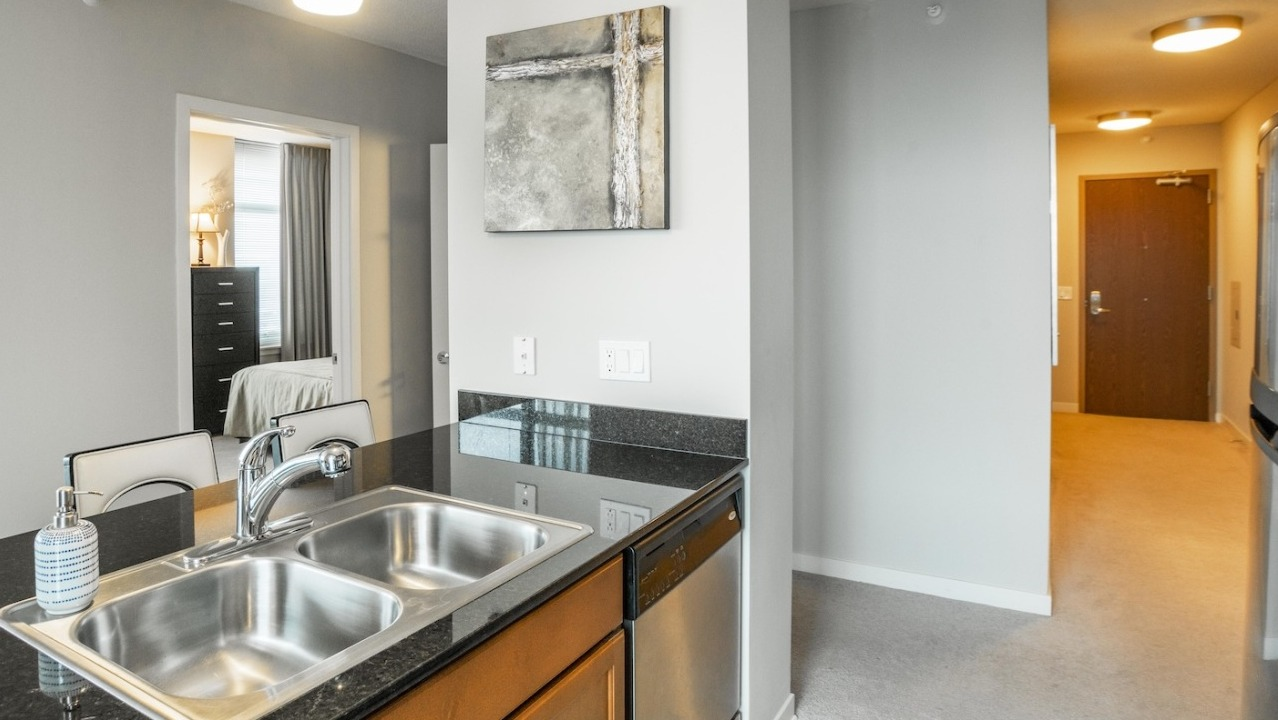 Open kitchen at Alta at K Station with granite countertop island, stainless sink, modern lighting, and hallway view leading to the entry