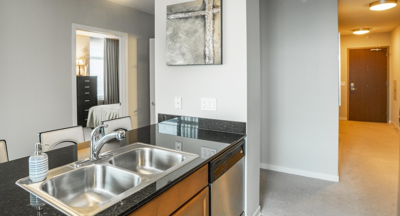 Open kitchen at Alta at K Station with granite countertop island, stainless sink, modern lighting, and hallway view leading to the entry