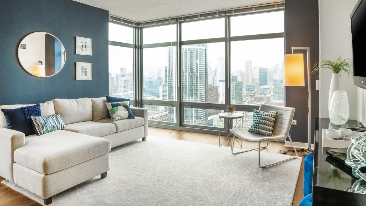 Bright living room at Alta at K Station with floor-to-ceiling windows, sectional sofa, wall-mounted TV, and sweeping Chicago skyline views