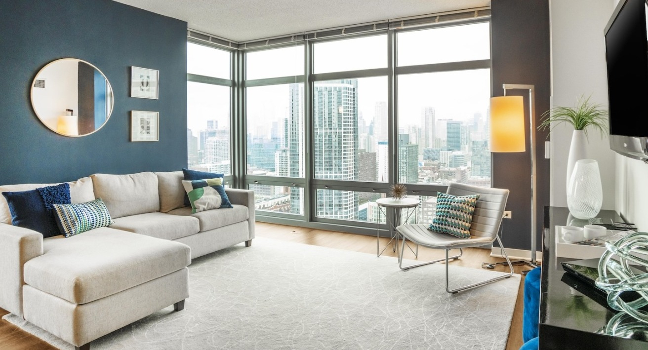 Bright living room at Alta at K Station with floor-to-ceiling windows, sectional sofa, wall-mounted TV, and sweeping Chicago skyline views