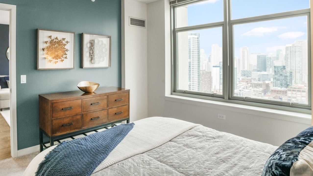 Bright master bedroom at Alta at K Station featuring large windows, dresser, blue accent wall, and modern art décor overlooking Chicago skyline