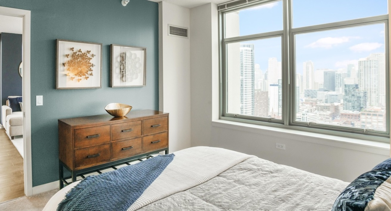 Bright master bedroom at Alta at K Station featuring large windows, dresser, blue accent wall, and modern art décor overlooking Chicago skyline