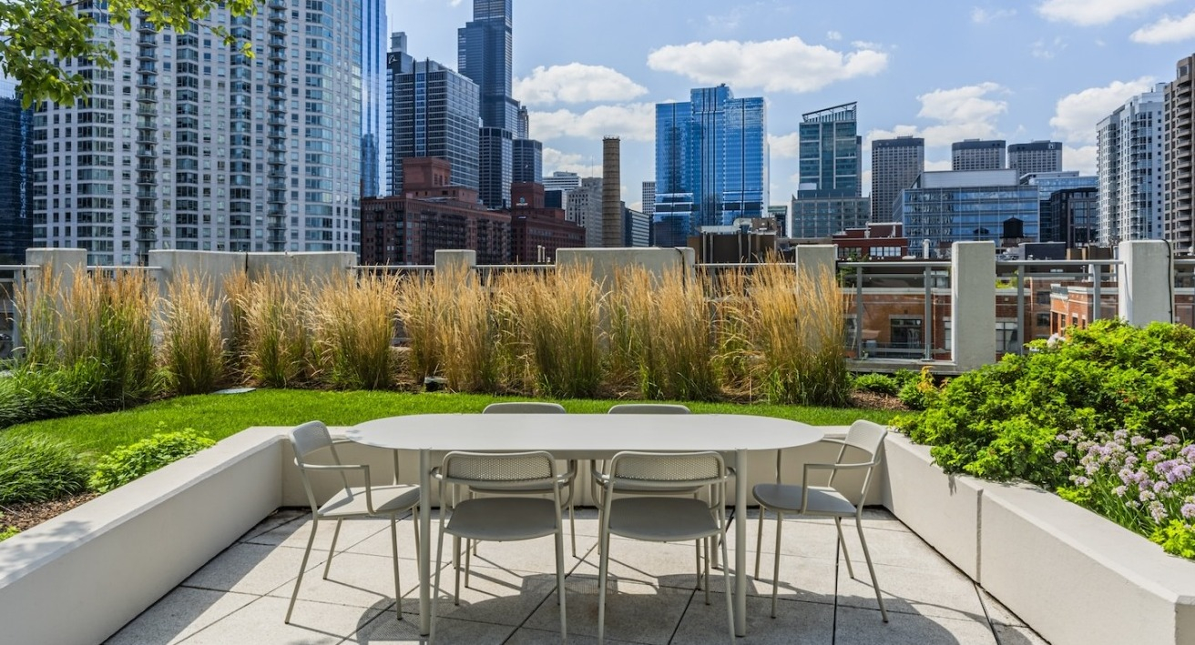 Outdoor dining area with table and chairs on the rooftop at Alta at K Station, surrounded by greenery and views of downtown Chicago skyscrapers
