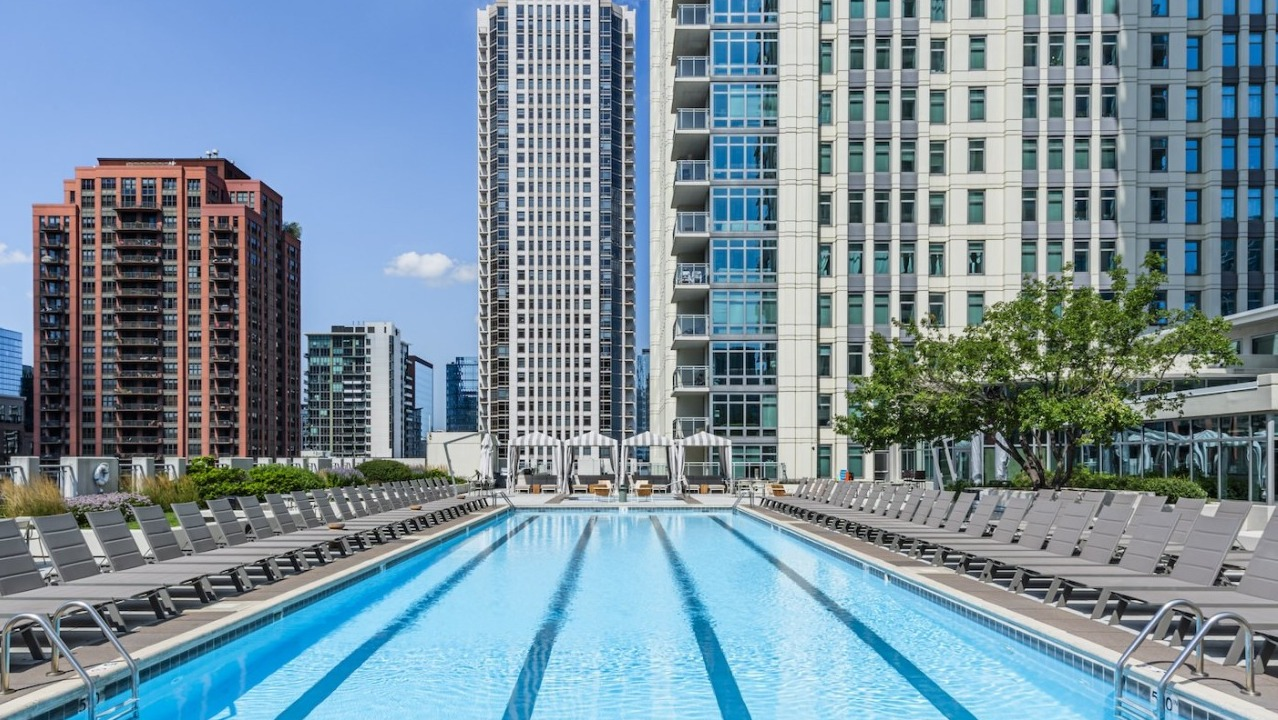 Rooftop swimming pool at Alta at K Station surrounded by lounge chairs, with panoramic views of Chicago high-rises and blue summer skies