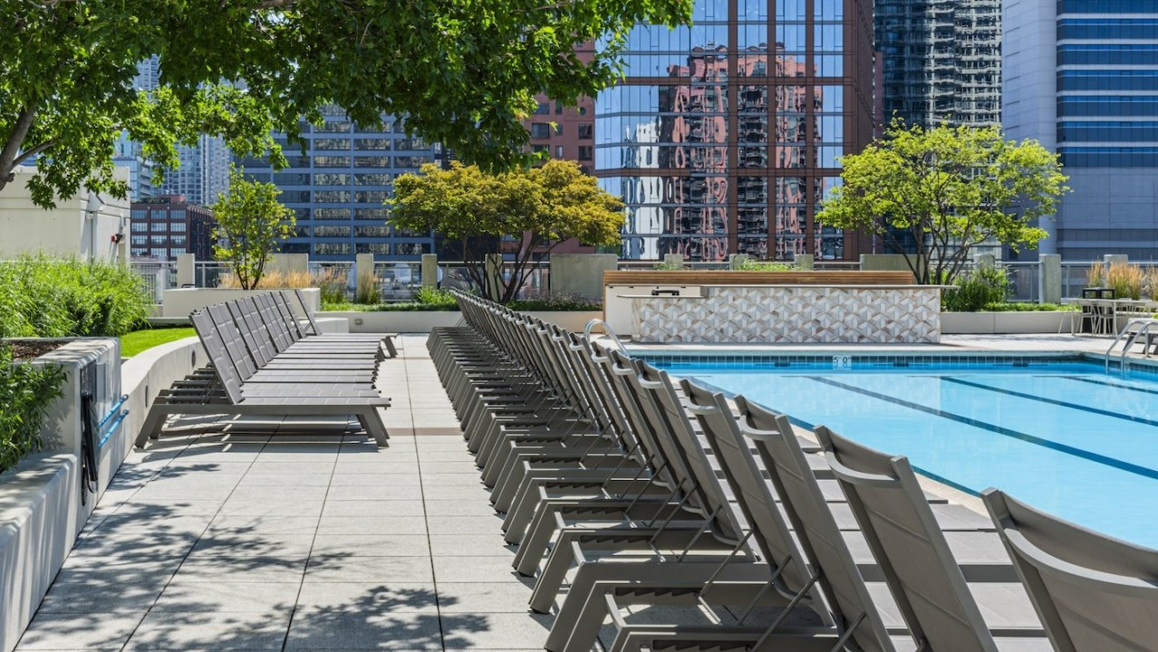 Rooftop pool deck at Alta at K Station with rows of lounge chairs shaded by trees, set against Chicago’s modern high-rise skyline
