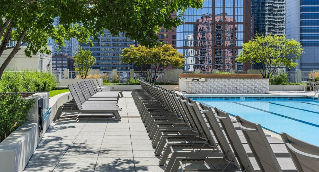 Rooftop pool deck at Alta at K Station with rows of lounge chairs shaded by trees, set against Chicago’s modern high-rise skyline